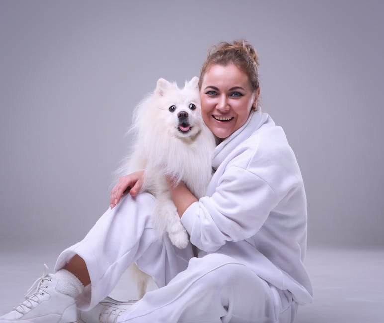 Smiling woman in white attire hugs her fluffy dog against a gray studio backdrop.