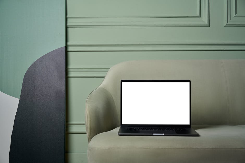 Modern netbook with white screen placed on soft couch near green uneven wall and big painting in light room